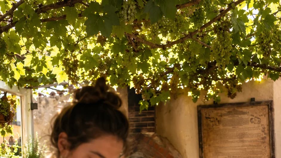 Solarpunk bakery under a grape trellis; baker shapes sourdough on a marble bench beside a wood-fired brick oven